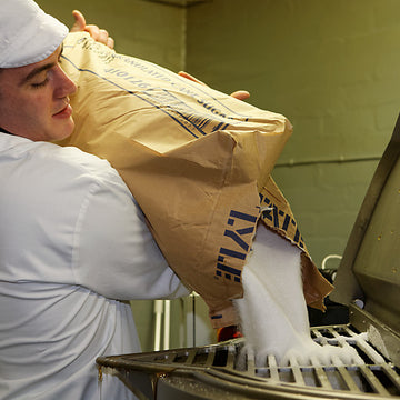 A man in a white overall and cap pouring a big bag of sugar into the opening of a food mixing vessel