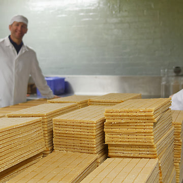 Two workers stood up wearing white overalls and caps are facing each other. In the foreground is stacks of Brown Kendal Mint Cake that are rectangular in shape
